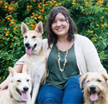Bio photo of Ginny Sims, smiling in a white sweater over a green top withi three large white dogs